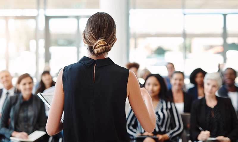 The back of a female public speaker presenting in front of a group of people